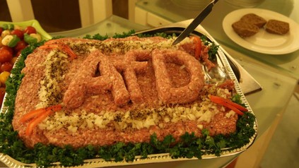 Auf einem Tablett befindet sich eine große Portion Mett aus dem das Wort AfD geformt wurde. Eine Person beginnt gerade damit das Mett anzuschneiden