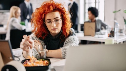 Junge FRau isst einen Snack im Büro vor ihrem Bildschirm