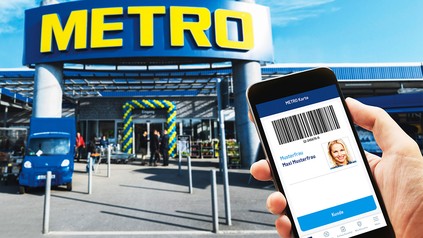 Metro Großhandelsmarkt im Hintergrund, im Vordergrund eine Hand, die ein Smartphone hält mit der geöffneten Metro Companion App