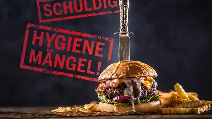 Ein Hamburger, in dem ein Messer steckt. An der Wand dahinter steht "schuldig!"